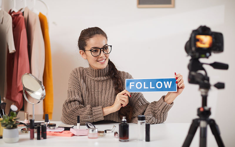 Portrait of contemporary young woman holding FOLLOW word while filming video for beauty and lifestyle channel, copy space